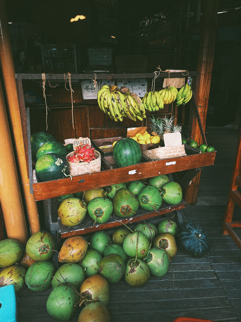 A rustic market stall filled with tropical fruits including green coconuts, bananas, pineapples, watermelon, and dragon fruit, showcasing fresh local produce and a natural, organic lifestyle aesthetic.