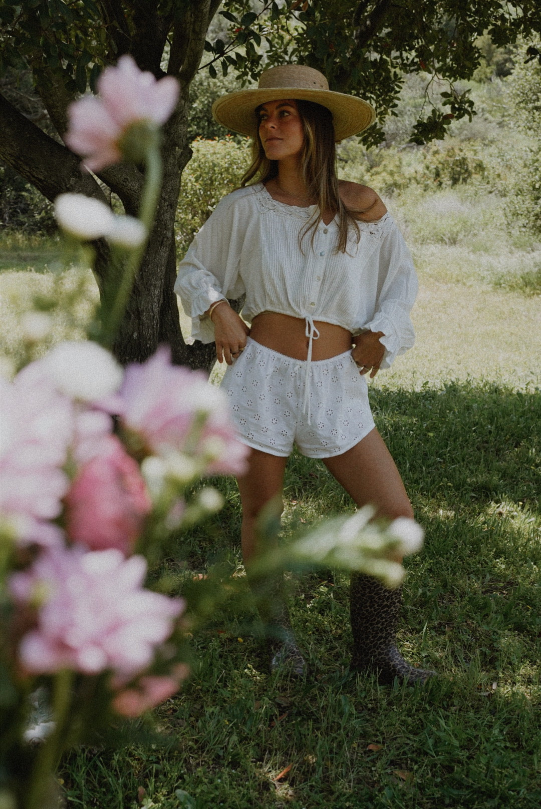 Woman standing under a tree wearing an airy, natural cotton outfit and straw hat, surrounded by flowers and greenery. A visual representation of breathable, skin-friendly fabrics supporting holistic skincare.