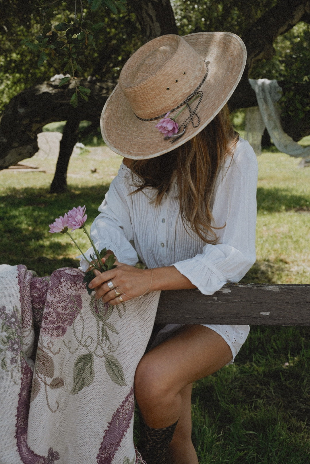 Mom in a wide-brimmed straw hat sitting quietly outdoors, holding fresh flowers and a vintage-style blanket—embracing a slow, restorative moment of self-care in nature.